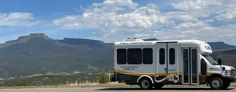 SCCOG bus parked on a hill with a picturesque view of Fishers peak and a blue sky with white hanging clouds in the background.
